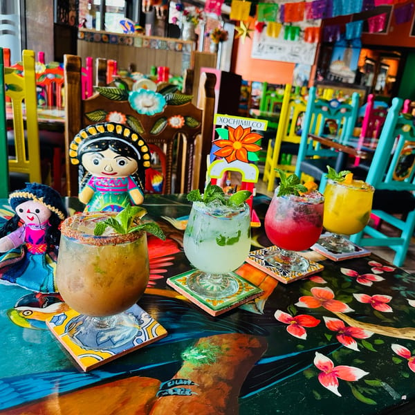 Four colorful Mexican cocktails with mint and fruit garnishes on a vibrant, painted table with dolls.