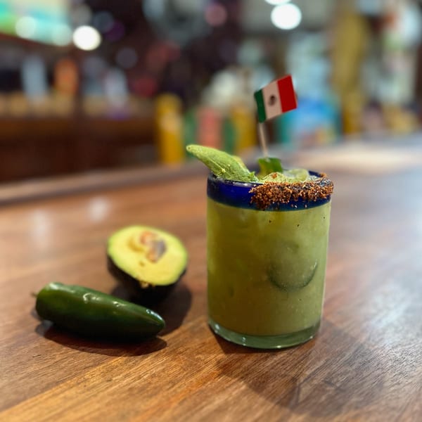 Avocado margarita with a Mexican flag, avocado half, and jalapeño on a wooden bar.