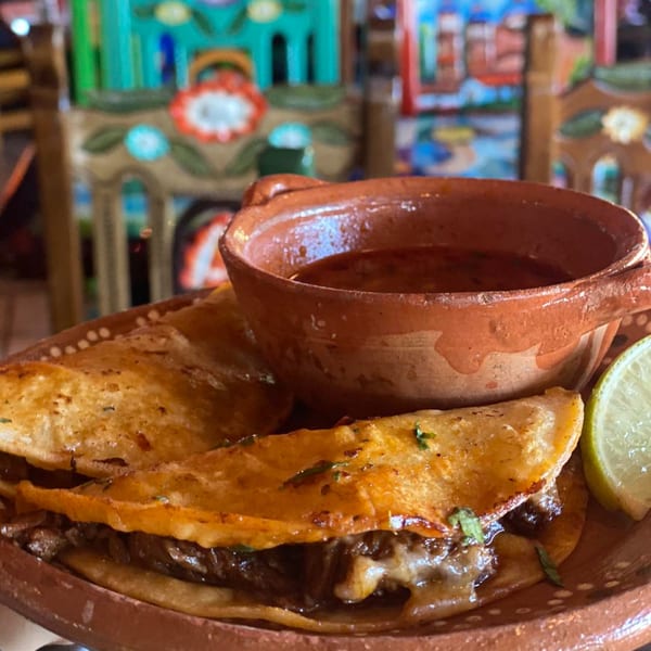 Three golden-brown birria tacos with melted cheese and meat, a side of consommé, and a lime wedge.