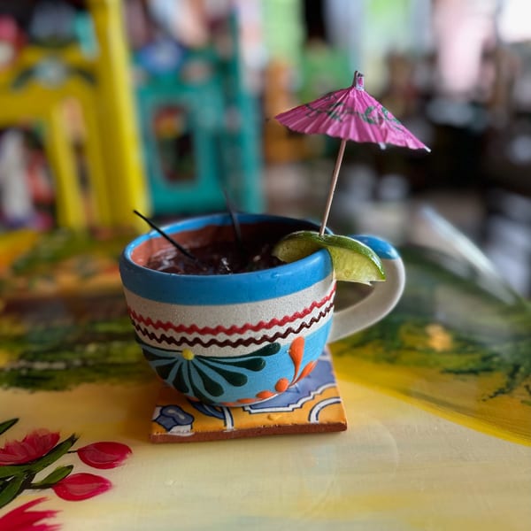 A colorful Mexican-style mug filled with a dark drink, garnished with a lime and pink umbrella.