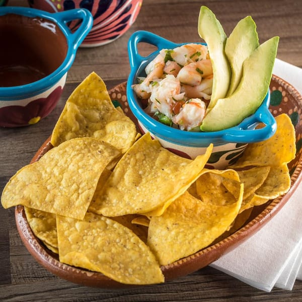 Shrimp ceviche with avocado slices in a blue bowl, surrounded by tortilla chips on a rustic plate.