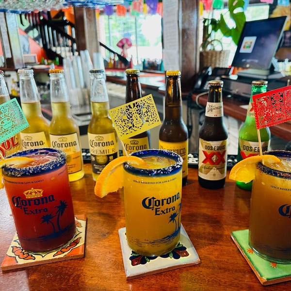 Three Corona Extra drinks with orange slices and festive picks, surrounded by various Mexican beer bottles at a bar.